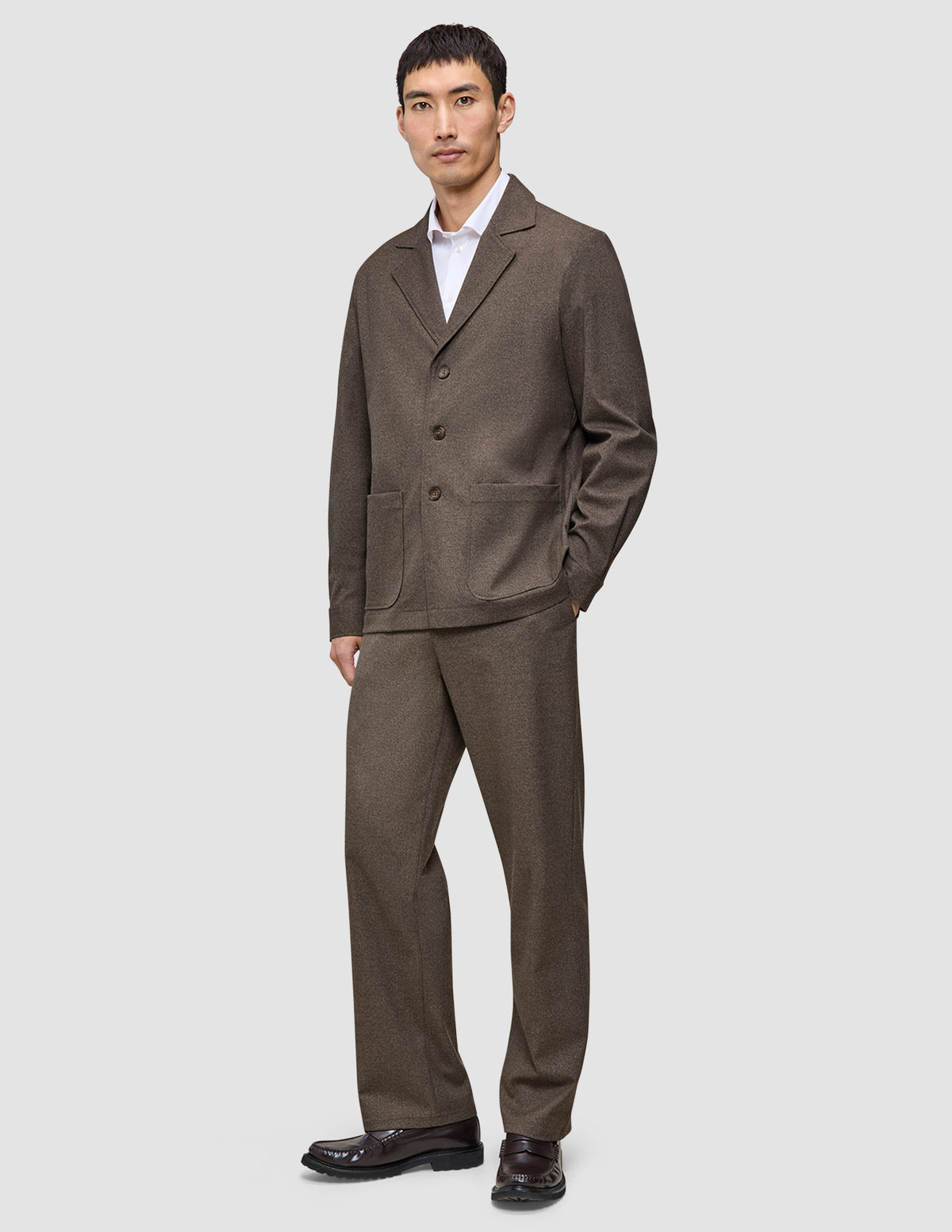 En mand iført en Atelier Overshirt i mørk beige og matchende bukser over en hvid skjorte med krave.