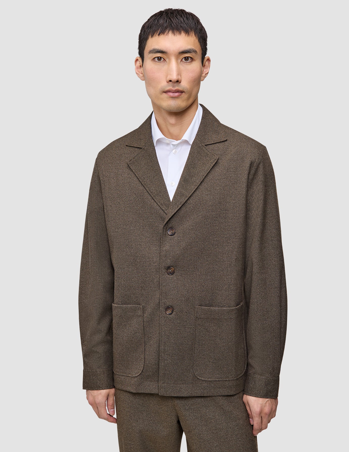 En mand har en brun Atelier Overshirt Dark Beige med udskåret krave og to påsatte lommer på over en hvid skjorte med krave.