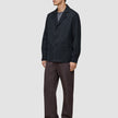 En mand iført en mørk marineblå Atelier Overshirt, en grå T-shirt, brune bukser og sorte loafers.
