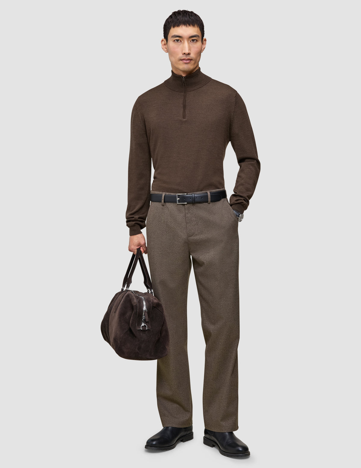 En mand iført en brun sweater med kvartlængde lynlås og et par Atelier Pants Relaxed i mørk beige, der holder en mørk taske.