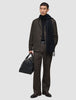En person iført en sort trøje, et tørklæde og et par mørkebrune Atelier Pants Relaxed, der holder en sort læderrejsetaske.