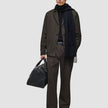 En person iført en sort trøje, et tørklæde og et par mørkebrune Atelier Pants Relaxed, der holder en sort læderrejsetaske.