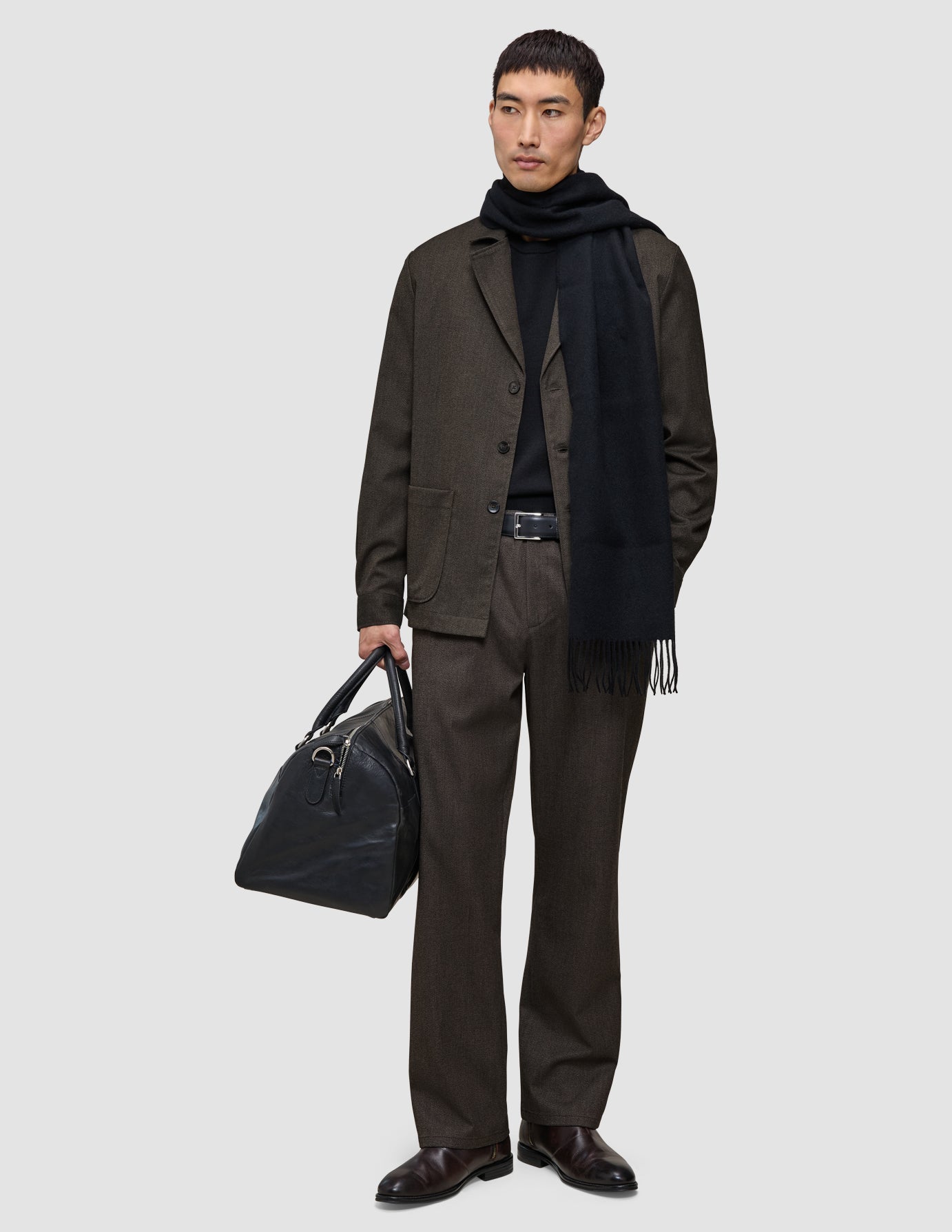 En person iført en sort trøje, et tørklæde og et par mørkebrune Atelier Pants Relaxed, der holder en sort læderrejsetaske.