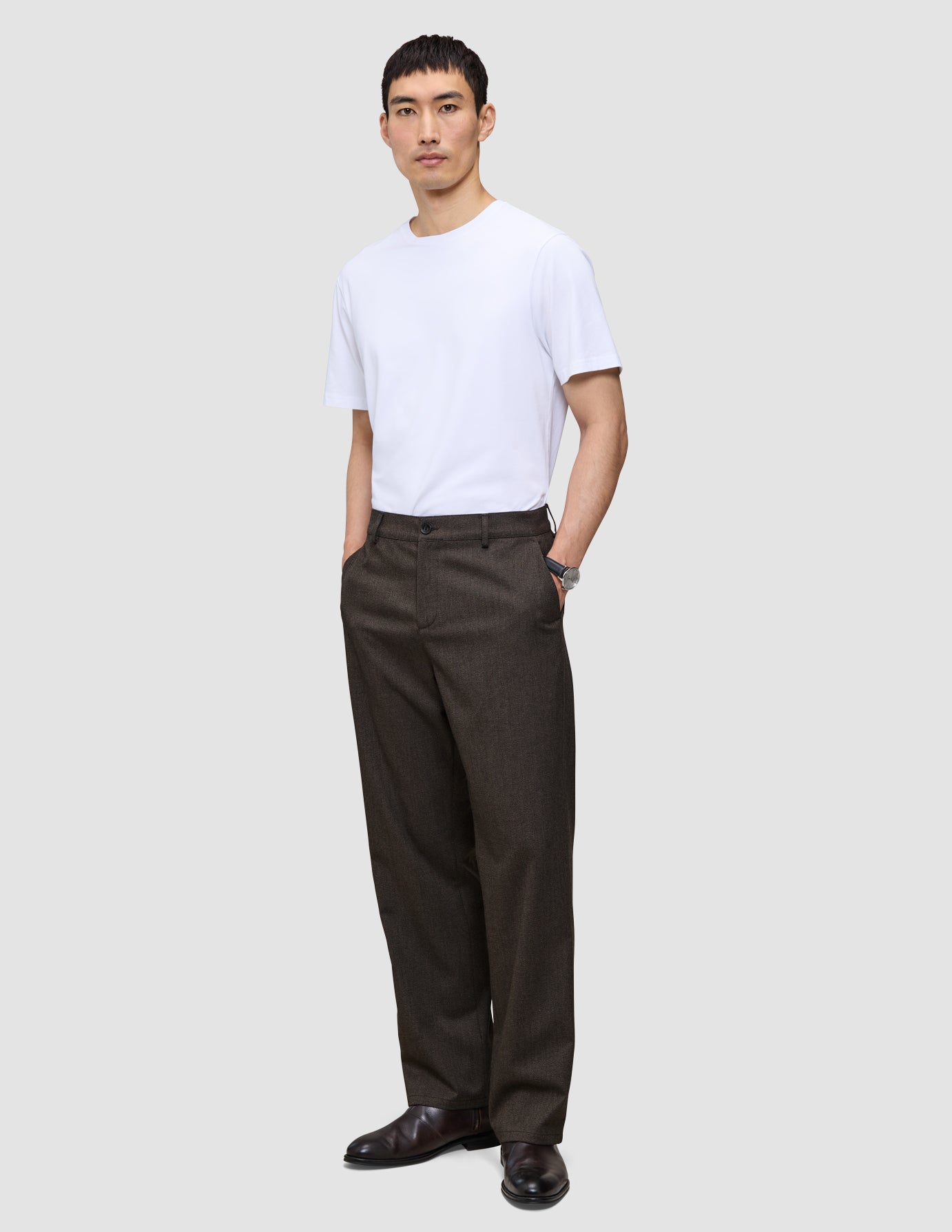 En mand har en hvid T-shirt på og et par Atelier Pants Relaxed i mørkebrun, syet i et afslappet-siddende brudt twill-stof.