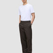 En mand har en hvid T-shirt på og et par Atelier Pants Relaxed i mørkebrun, syet i et afslappet-siddende brudt twill-stof.