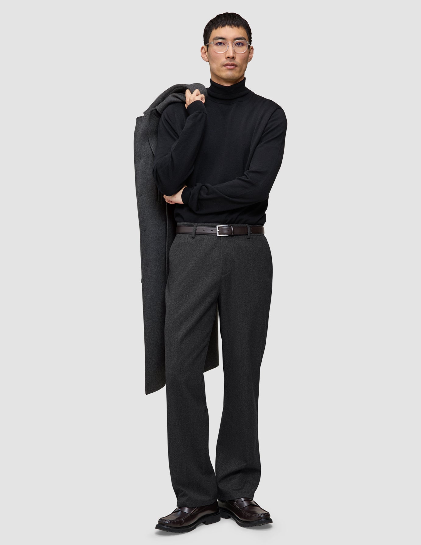 En person iført en sort rullekravebluse og et par mørkegrå Atelier Pants Relaxed med en frakke hængende over skulderen.