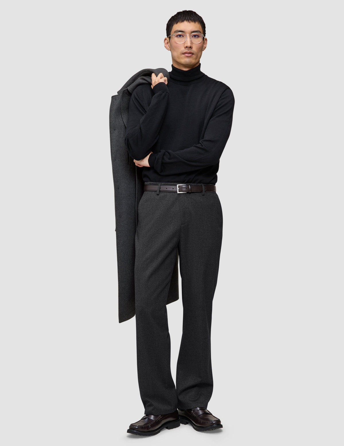 En person iført en sort rullekravebluse og et par mørkegrå Atelier Pants Relaxed med en frakke hængende over skulderen.