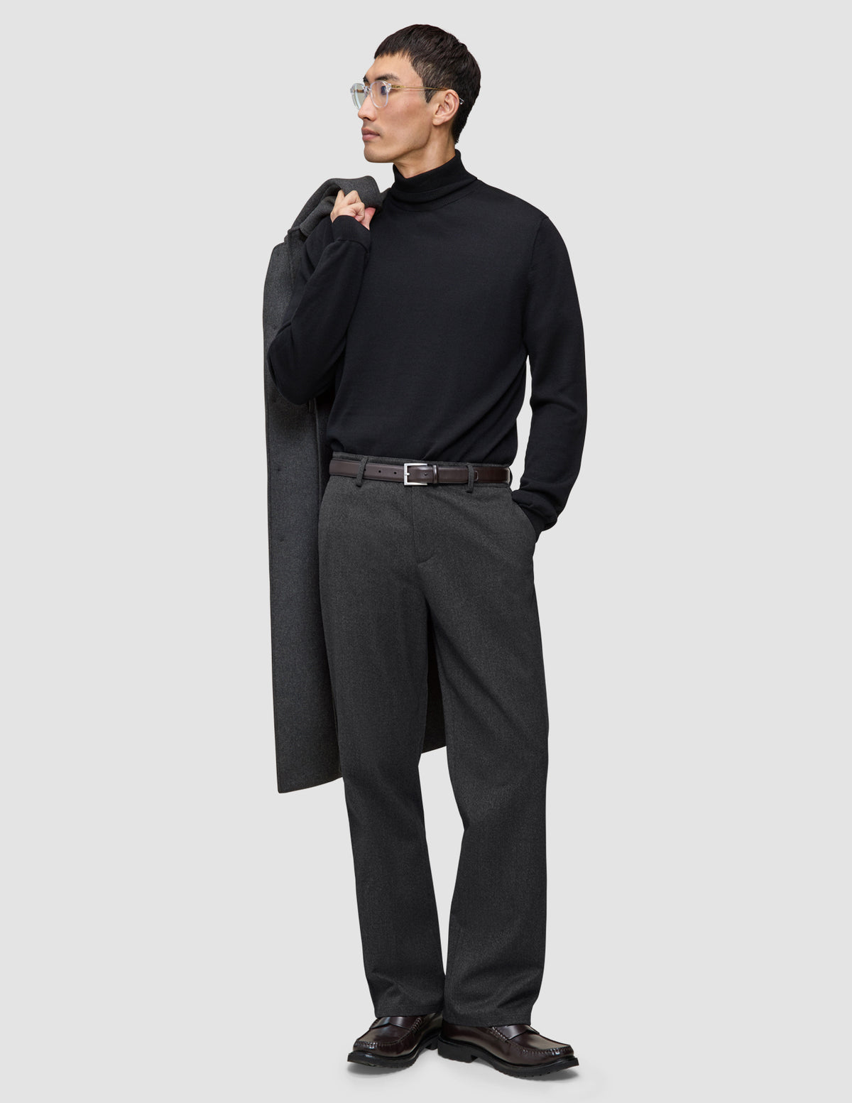 En mand i en sort rullekravebluse og mørkegrå Atelier Pants Relaxed, der har en frakke over skulderen.