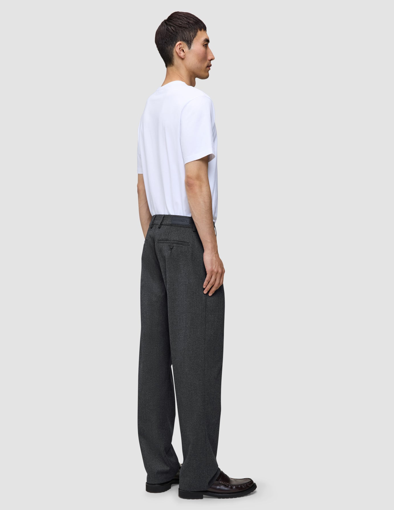 En mand i en hvid T-shirt, iført Atelier Pants Relaxed i mørkegrå og mørke loafers.