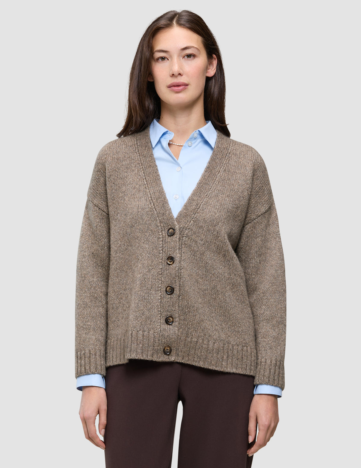 En person, der har en tyk, blød uldstrikket cardigan til kvinder i farven taupe melange på over en lyseblå skjorte med krave.