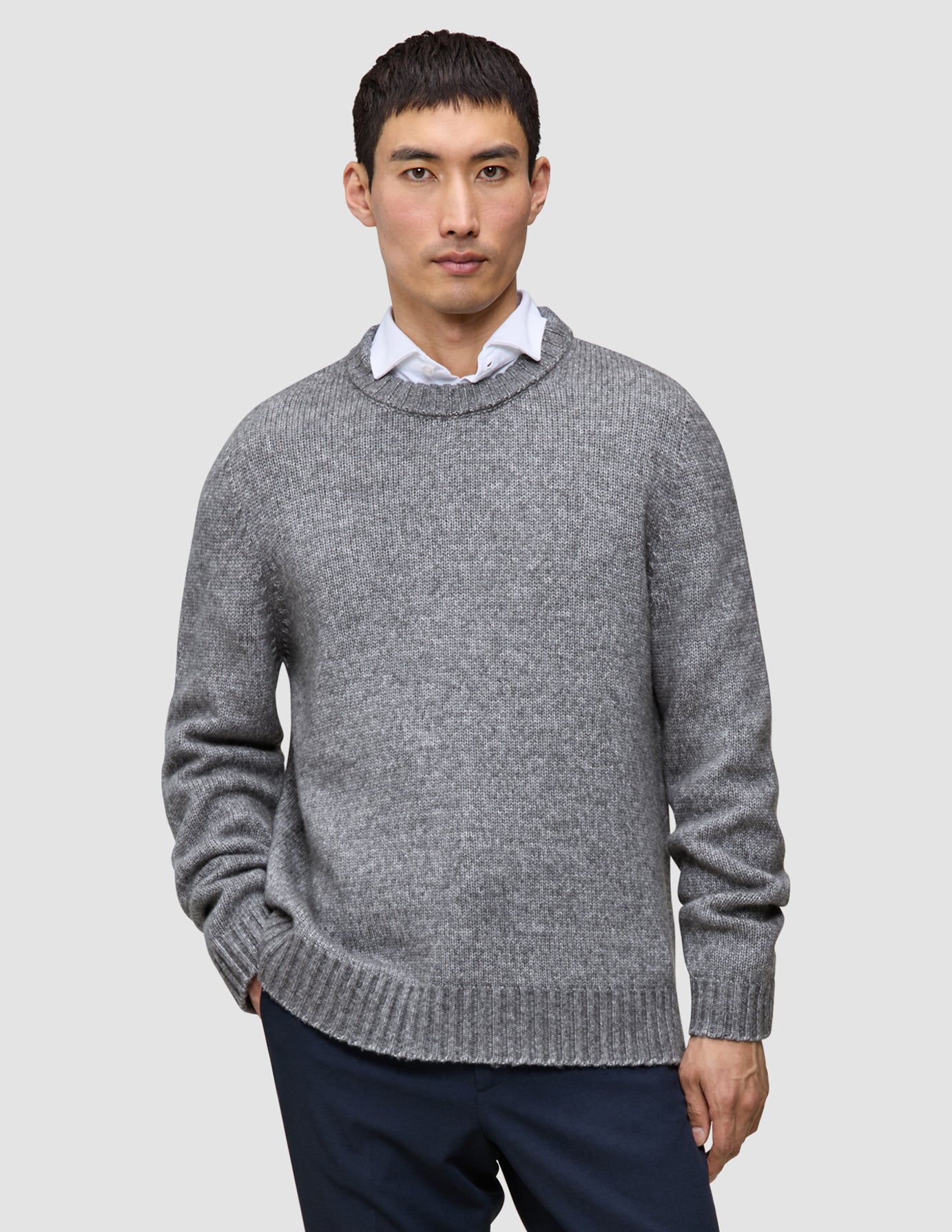 En person, der har en tyk, blød uldstrikket sweater med rund hals i farven Glacier Grey på over en hvid skjorte med krave.