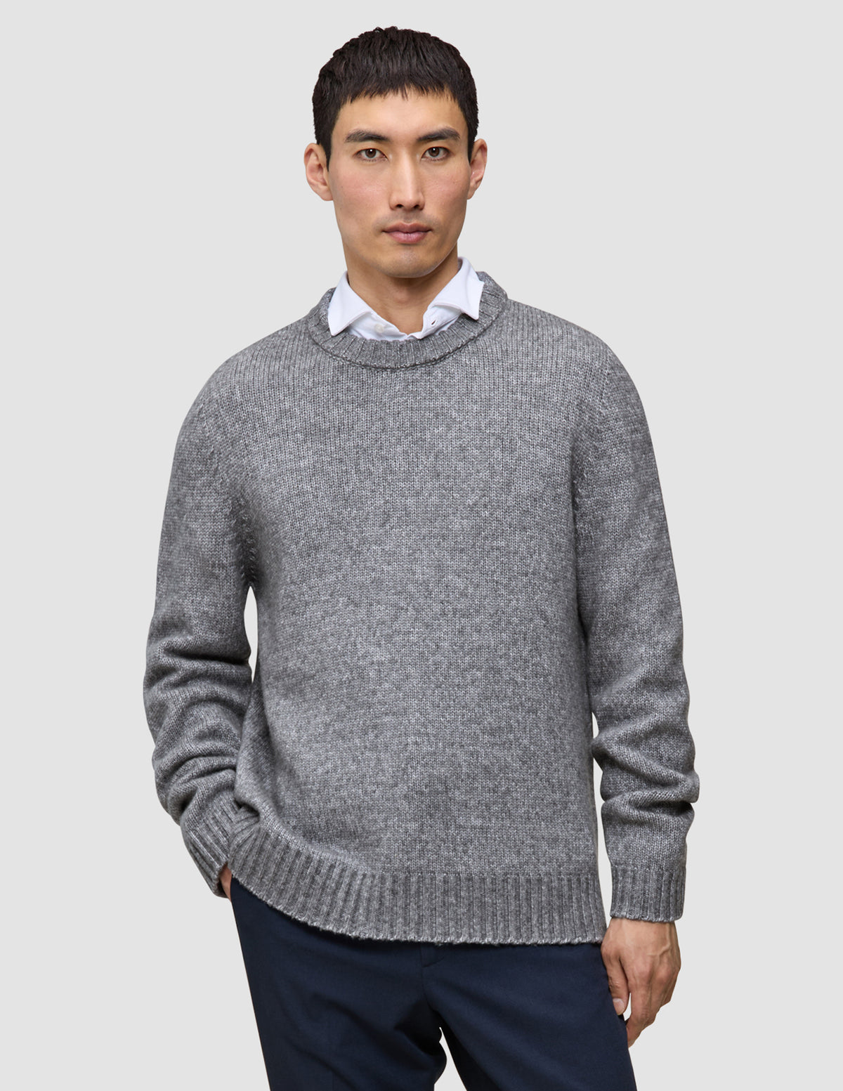 En person, der har en tyk, blød uldstrikket sweater med rund hals i farven Glacier Grey på over en hvid skjorte med krave.