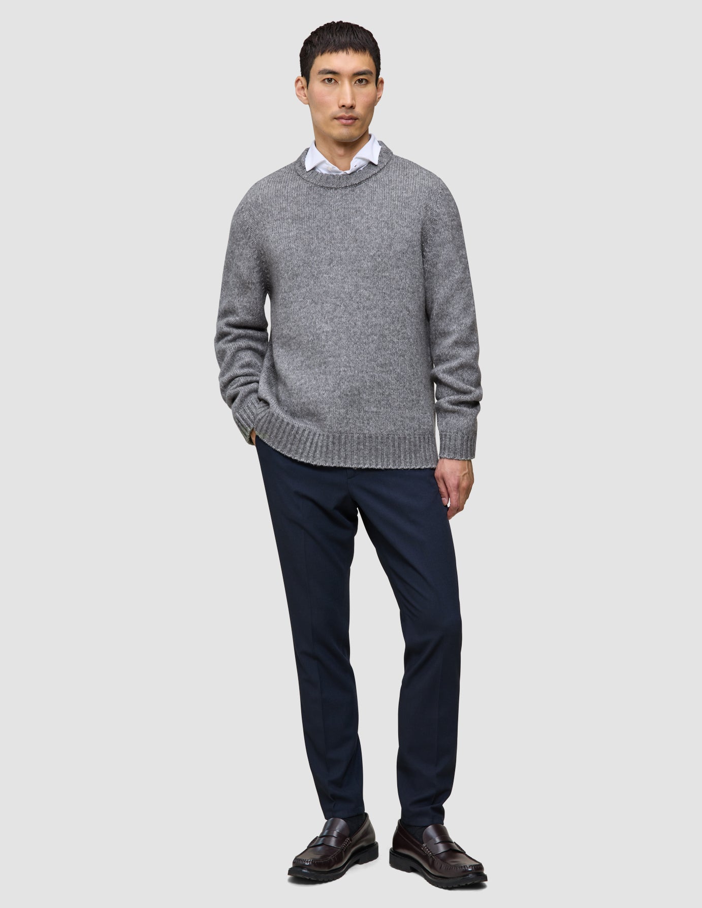 En mand iført en tyk, blød uldstrikket sweater med rund hals i farven Glacier Grey over en hvid skjorte med krave, marineblå bukser og loafers.