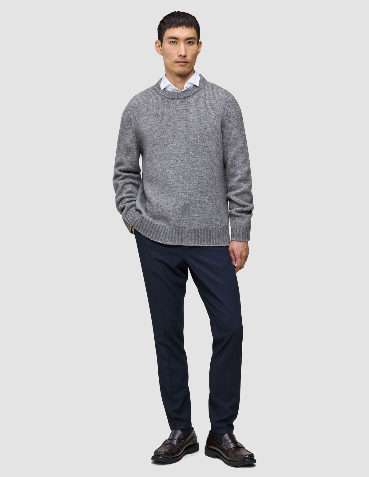 En mand iført en tyk, blød uldstrikket sweater med rund hals i farven Glacier Grey over en hvid skjorte med krave, marineblå bukser og loafers.
