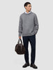 En mand iført en tyk, blød uldstrikket sweater med rund hals i farven Glacier Grey og mørke bukser bærer en brun rejsetaske.