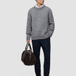 En mand iført en tyk, blød uldstrikket sweater med rund hals i farven Glacier Grey og mørke bukser bærer en brun rejsetaske.