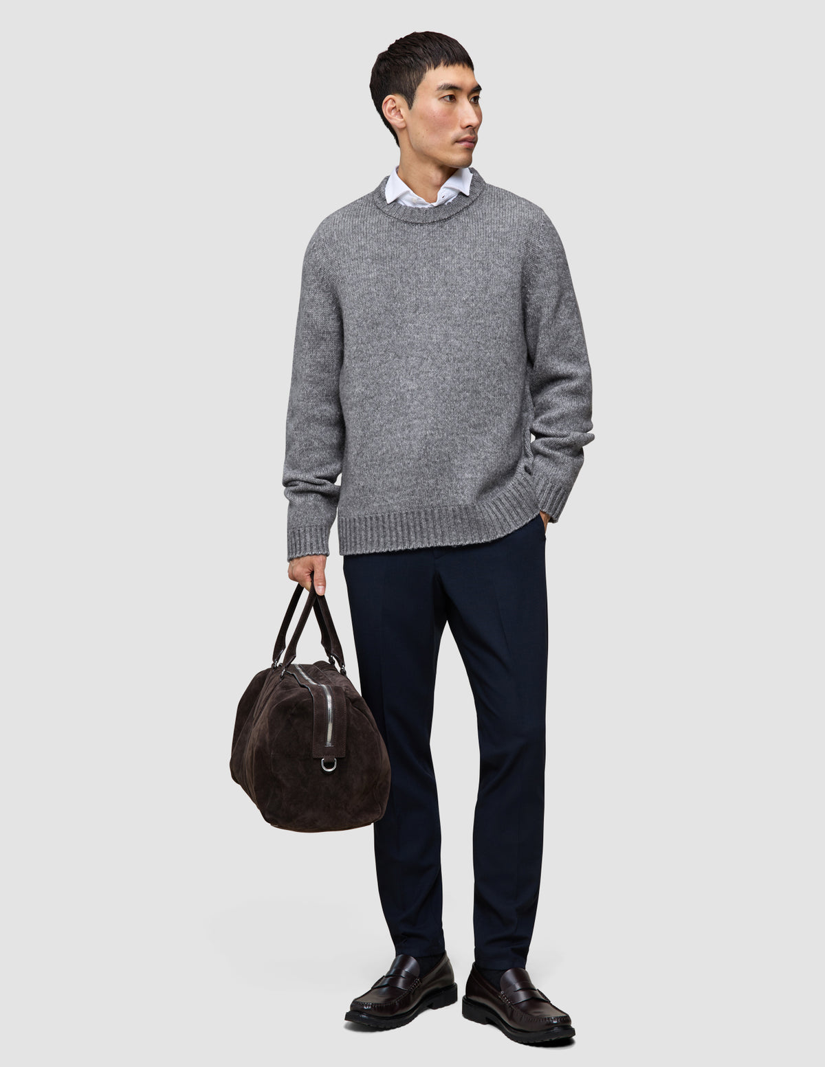 En mand iført en tyk, blød uldstrikket sweater med rund hals i farven Glacier Grey og mørke bukser bærer en brun rejsetaske.