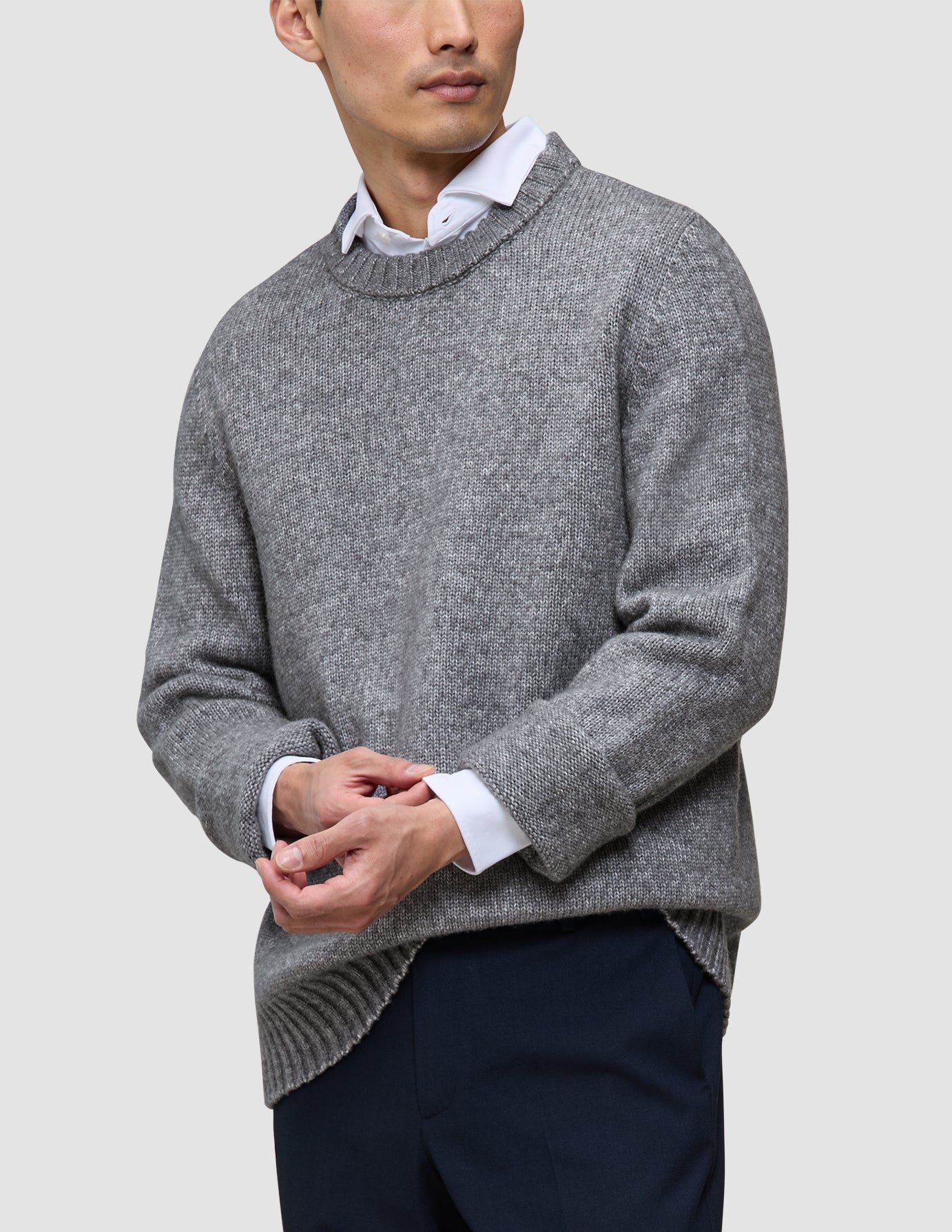 En person iført en tyk, blød uldstrikket sweater med rund hals i farven Glacier Grey over en hvid skjorte med krave og mørke bukser.