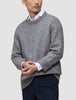 En person iført en tyk, blød uldstrikket sweater med rund hals i farven Glacier Grey over en hvid skjorte med krave og mørke bukser.
