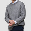En person iført en tyk, blød uldstrikket sweater med rund hals i farven Glacier Grey over en hvid skjorte med krave og mørke bukser.