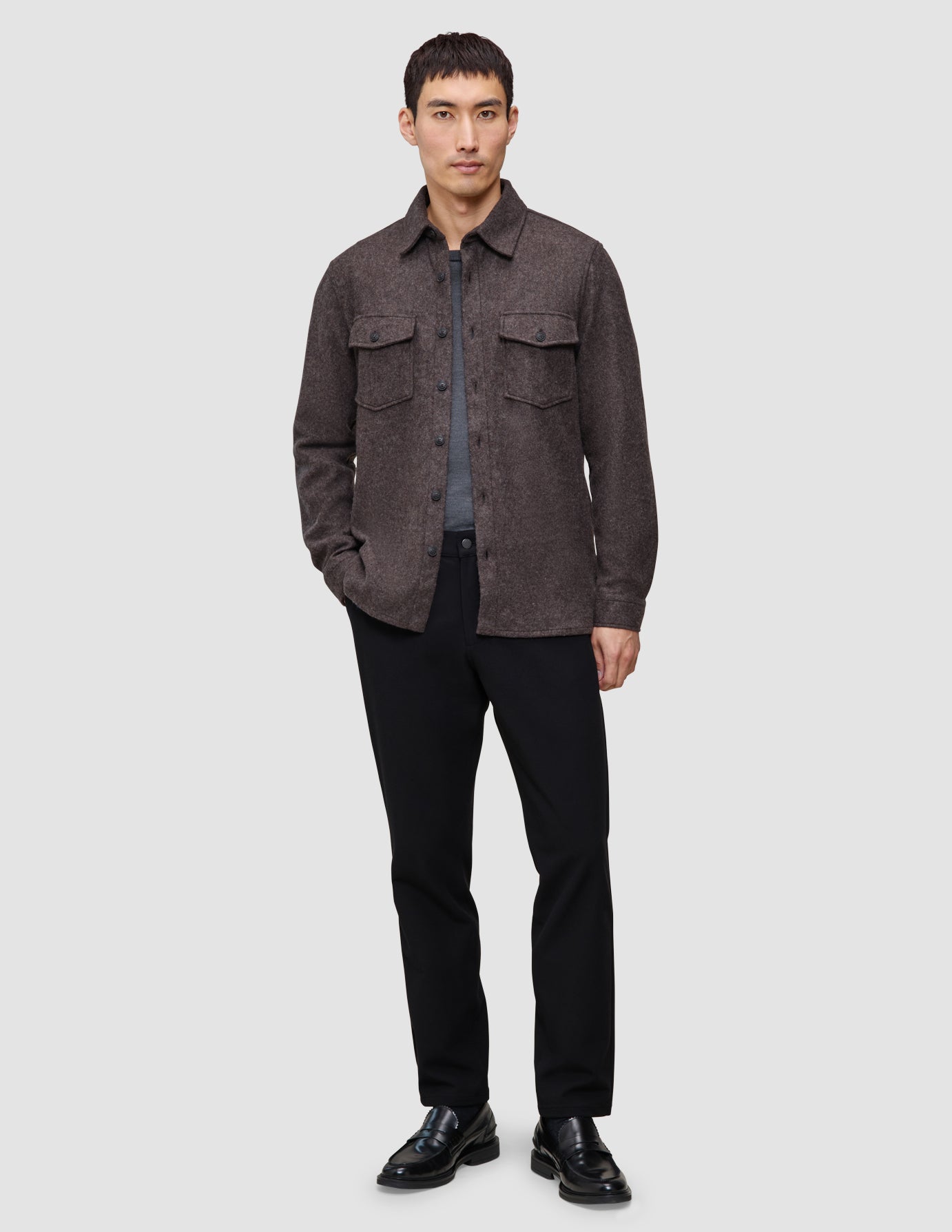 En mand iført en dobbeltlags uldskjorte i sadelbrun over en grå T-shirt, sorte bukser og sorte loafers.