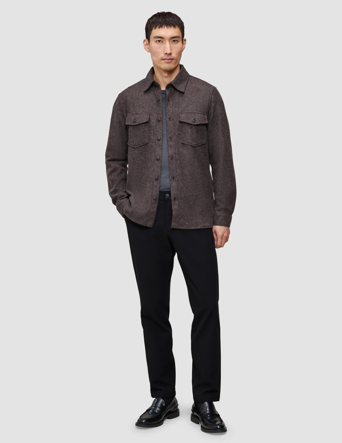 En mand iført en dobbeltlags uldskjorte i sadelbrun over en grå T-shirt, sorte bukser og sorte loafers.