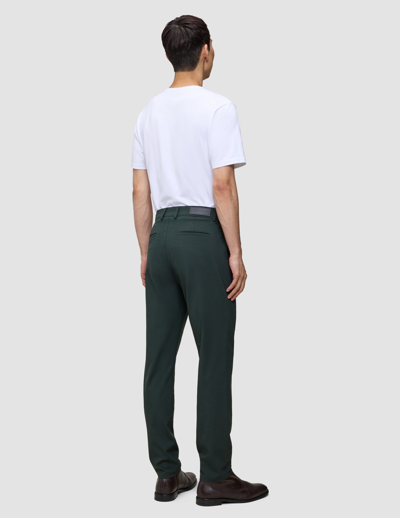 En person, set bagfra, har en hvid T-shirt på, Ease Pants Regular Northern Pine og brune lædersko.