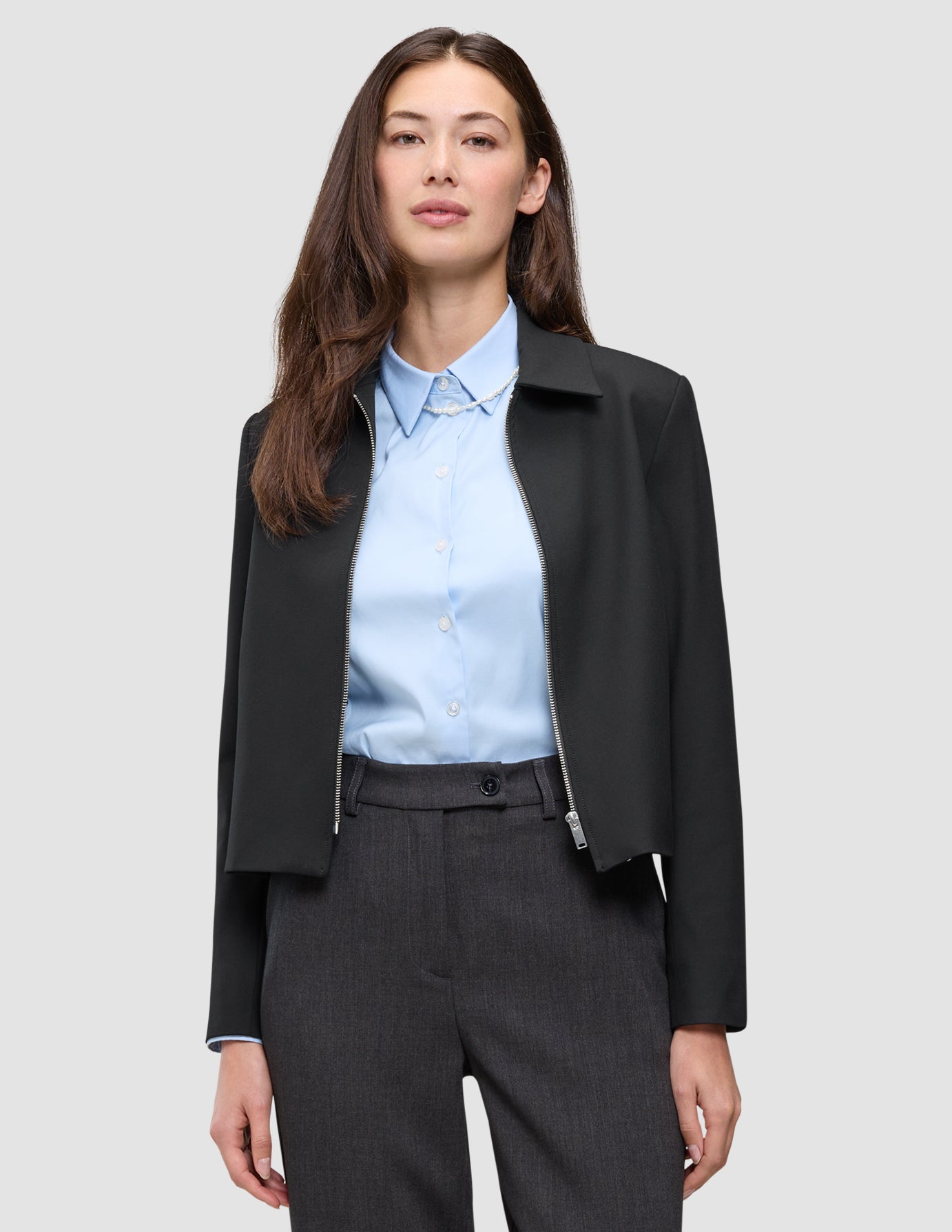 En person, der har en sort Essential Short Zip Jacket på over en lyseblå skjorte med knapper og grå bukser.