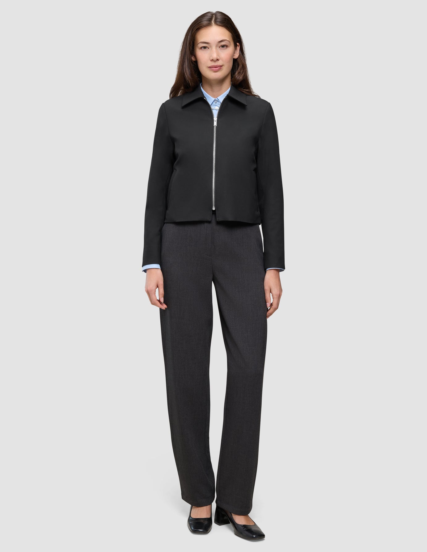 En kvinde iført en sort Essential Short Zip Jacket over en lyseblå skjorte med krave og mørke bukser.