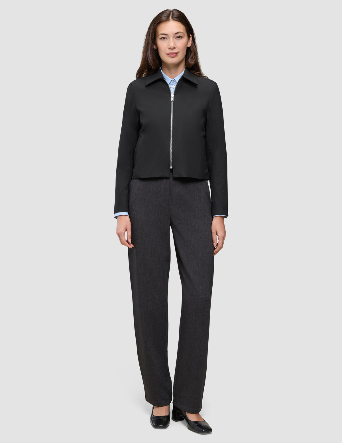 En kvinde iført en sort Essential Short Zip Jacket over en lyseblå skjorte med krave og mørke bukser.