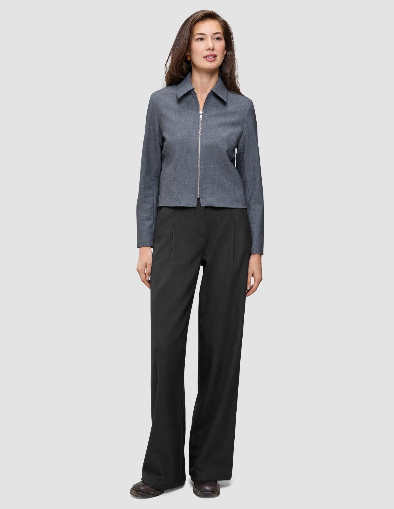 En kvinde iført en Essential Short Zip Jacket i mørkegrå melange og sorte bukser med brede ben.