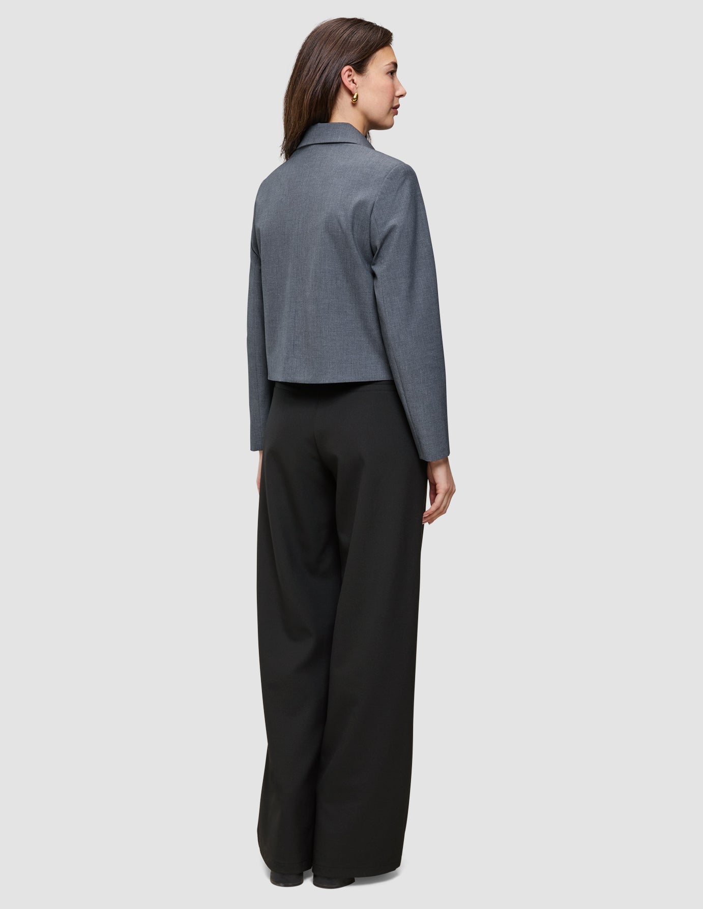 En person set bagfra, iført en Essential Short Zip Jacket i mørkegrå melange og sorte bukser med brede ben.