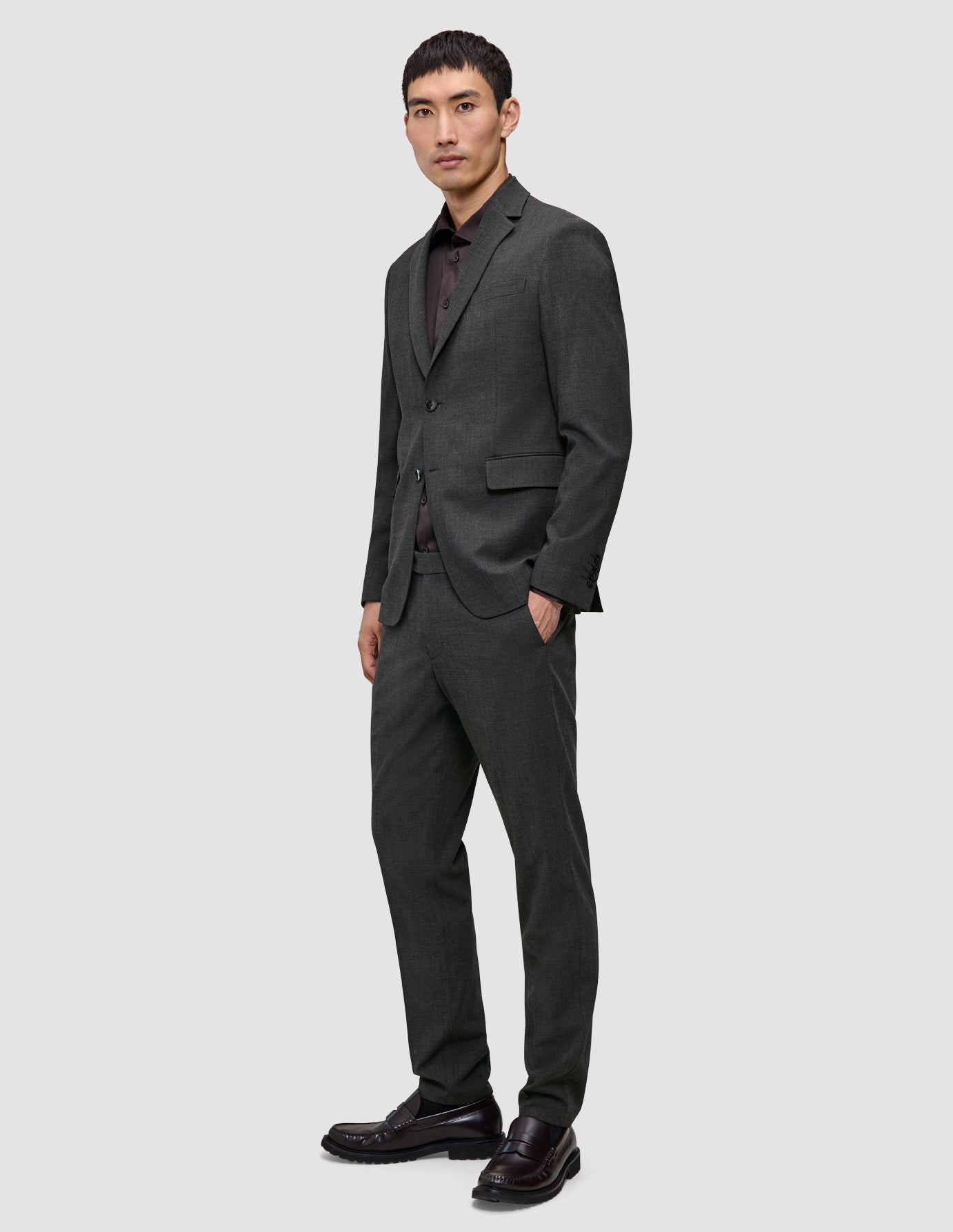 En mand iført en Motion Blazer Slim i farven Pepper Grey og matchende bukser over en mørk skjorte med knapper og loafers.