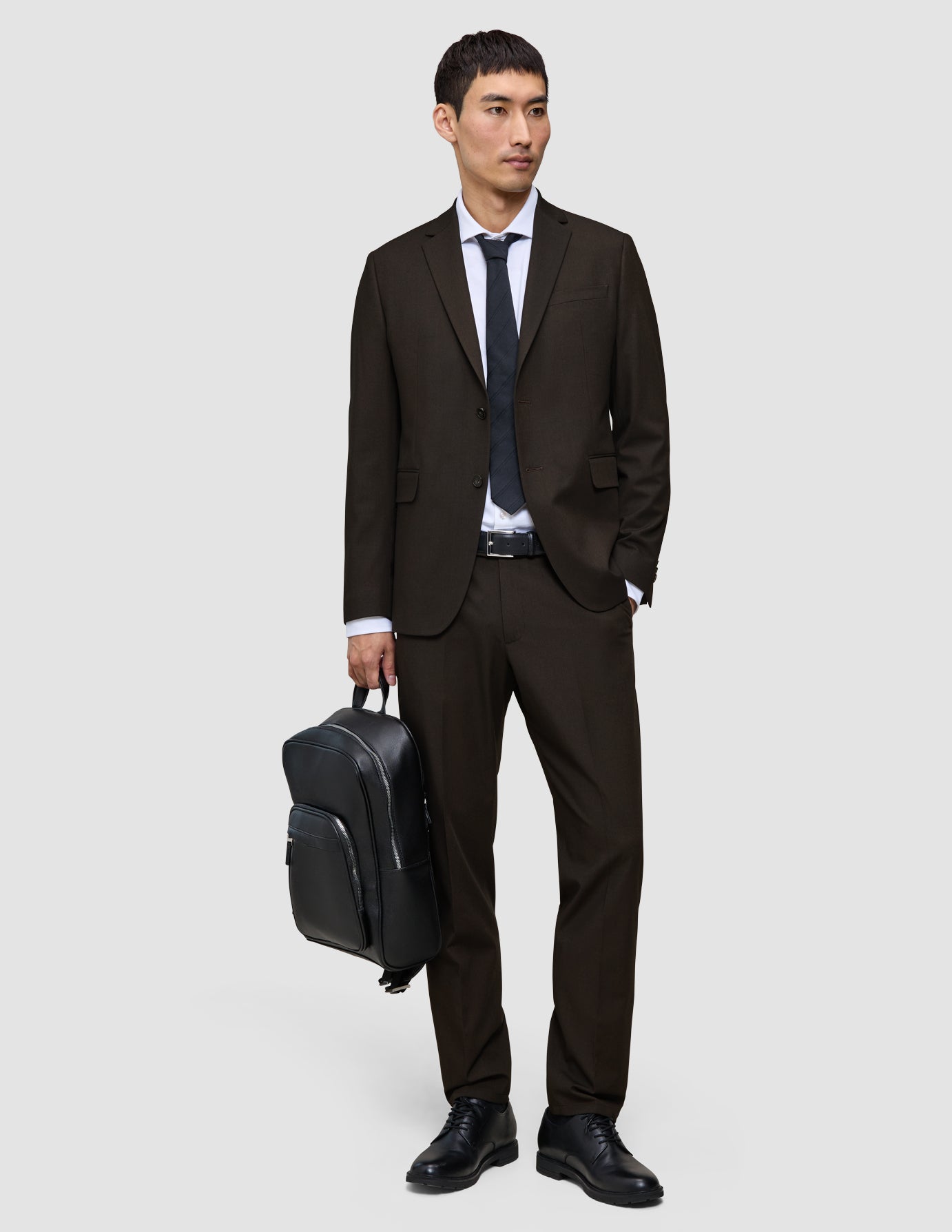 En mand iført et Motion Suit-jakkesæt i farven Regular Cognac, hvid skjorte og sort slips holder en sort rygsæk.