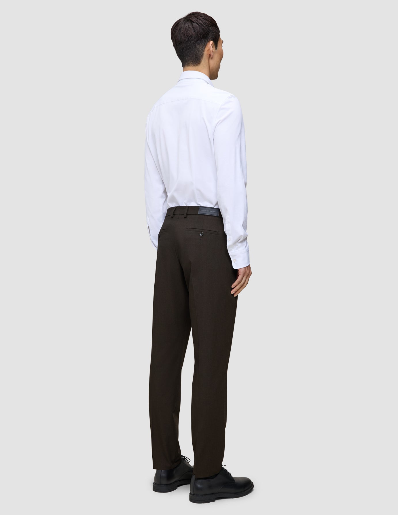 En person set bagfra, iført en hvid skjorte, Motion Suit-bukser i Regular-pasform i farven Cognac og sorte dressko.