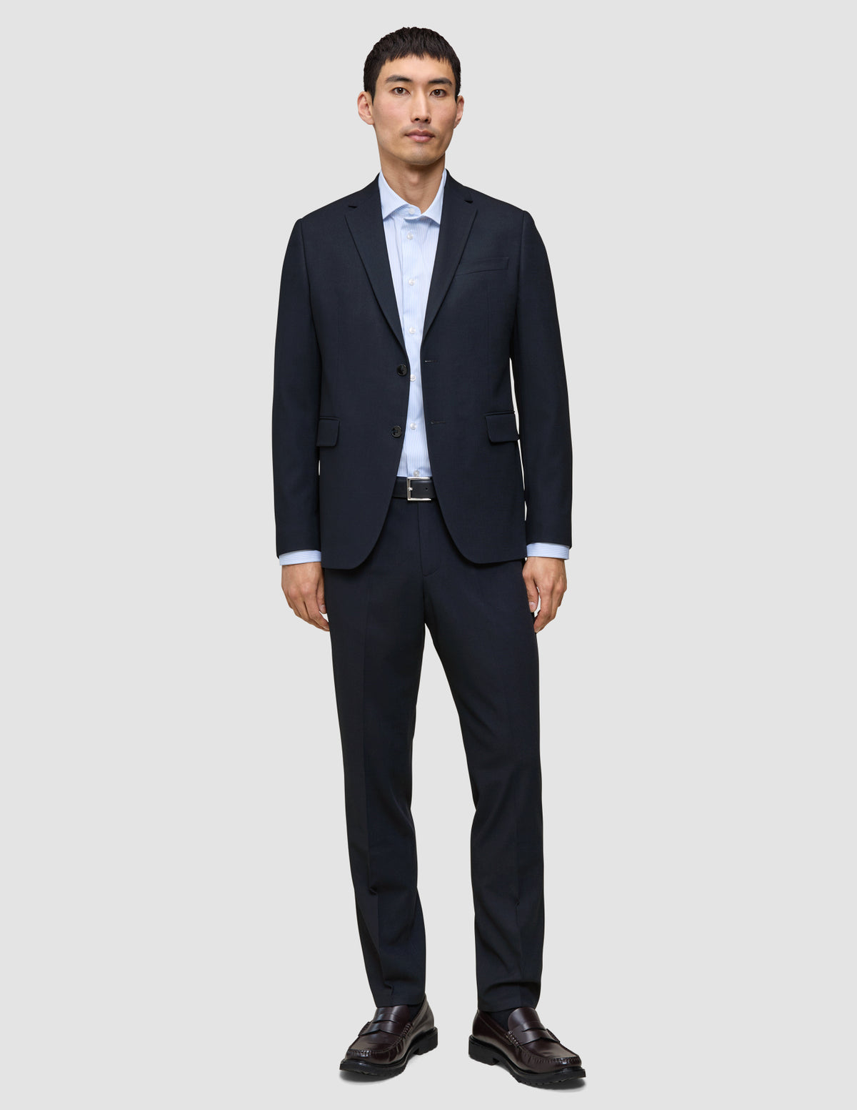 En mand iført Motion Suit Pants Regular Midnight Blue i et mørkeblåt jakkesæt, en lyseblå skjorte og mørke loafers.