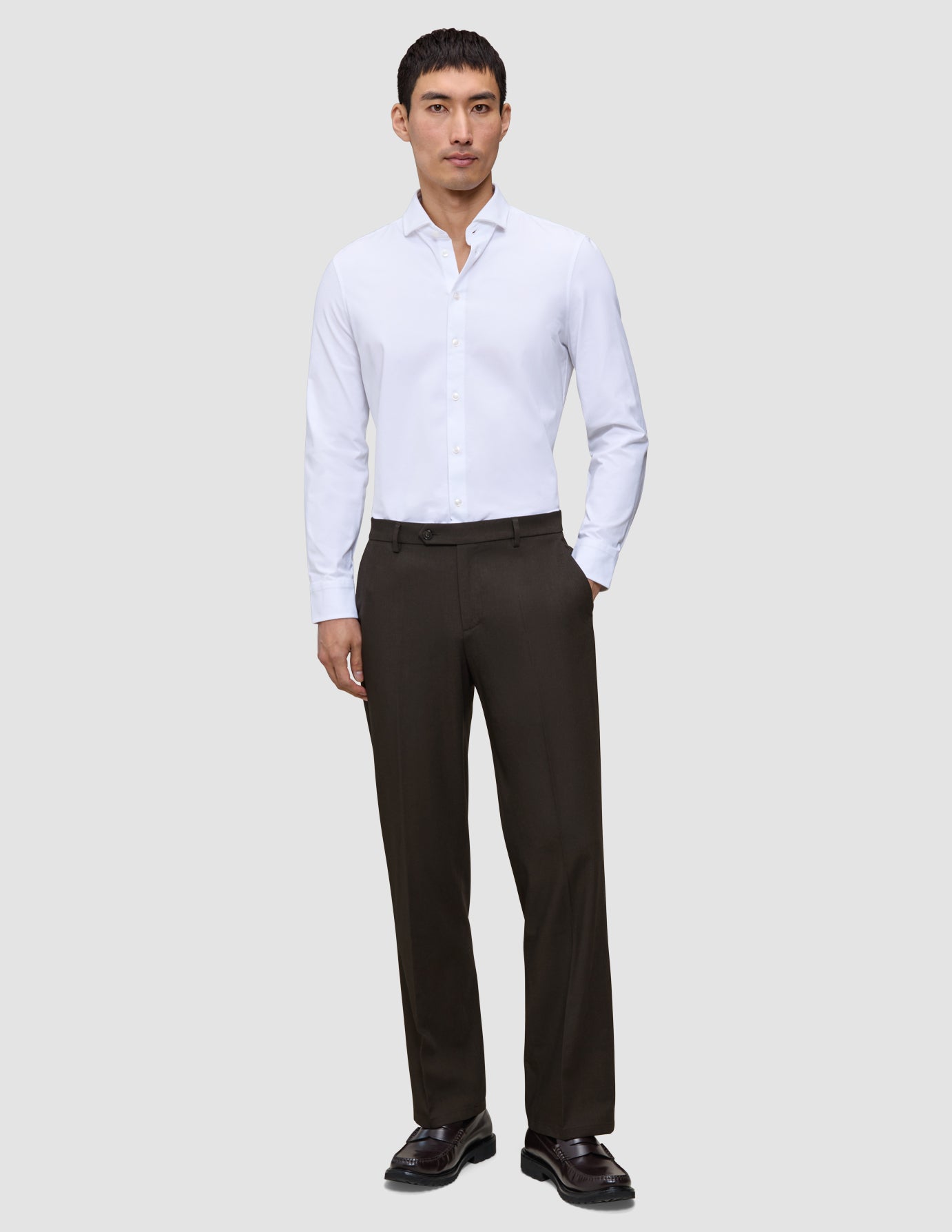 En mand iført en hvid skjorte med knapper, Motion Suit-bukser i afslappet pasform i farven Cognac og mørke loafers.
