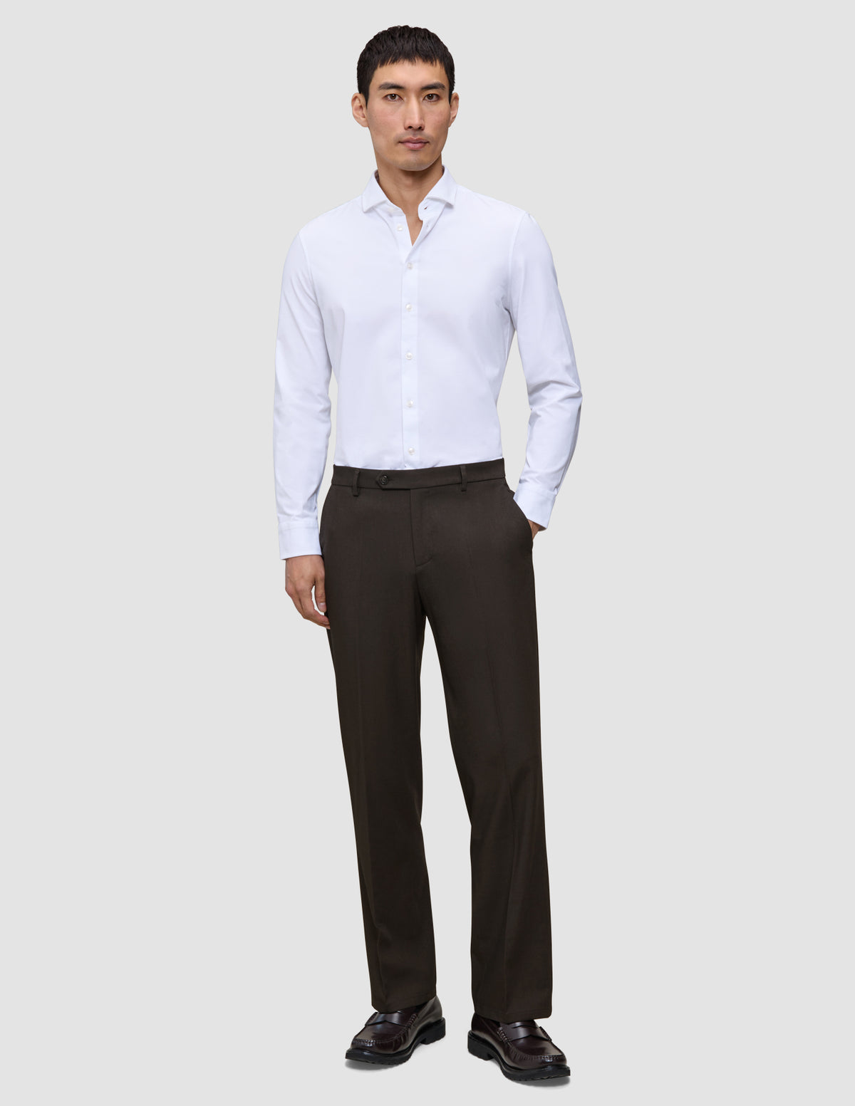 En mand iført en hvid skjorte med knapper, Motion Suit-bukser i afslappet pasform i farven Cognac og mørke loafers.