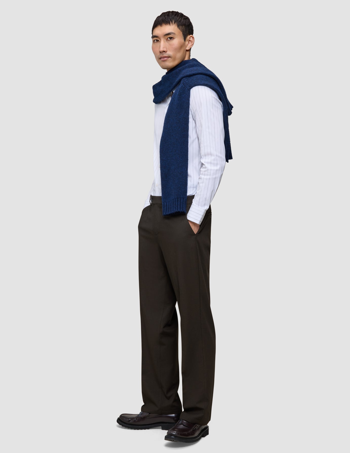 En mand i en hvid skjorte med tynde striber og en blå sweater har et par Motion Suit Pants Relaxed Cognac i mørkebrun på.
