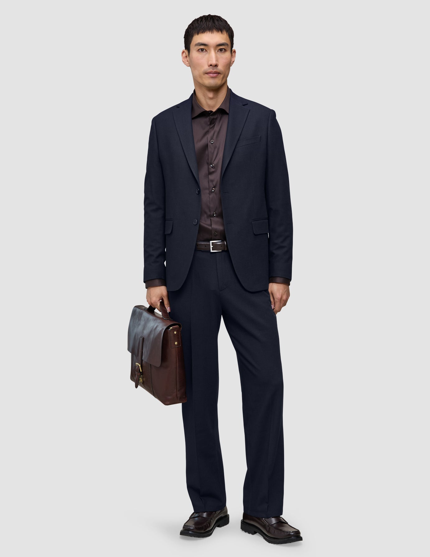 En mand iført et par marineblå Motion Suit-bukser i afslappet pasform Midnight Blue, en mørk skjorte og loafers holder en brun lædermappe.