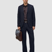 En mand iført et par marineblå Motion Suit-bukser i afslappet pasform Midnight Blue, en mørk skjorte og loafers holder en brun lædermappe.