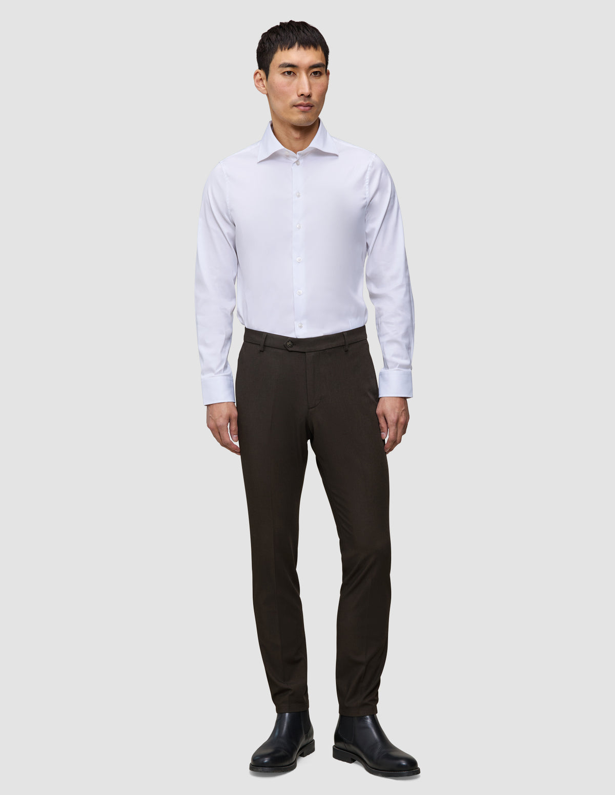 En mand iført en hvid skjorte med knapper, Motion Suit-bukser i Slim-pasform i farven Cognac og sorte støvler.
