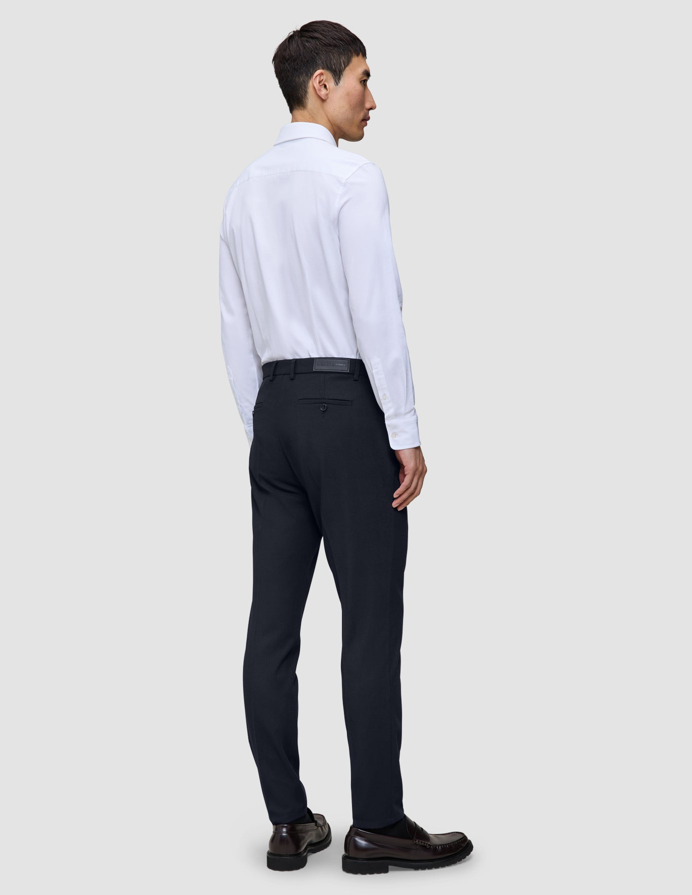En mand set bagfra iført en hvid skjorte med knapper, Motion Suit-bukser i slim fit i midnatsblå og mørke læderloafers.