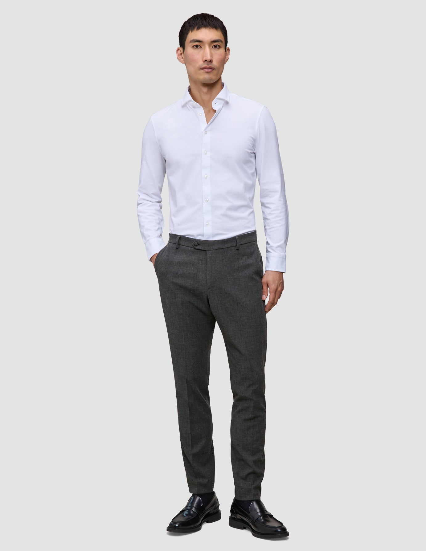 En mand i en hvid skjorte med knapper og et par Motion Suit-bukser i farven Slim Pepper Grey samt sorte loafers.