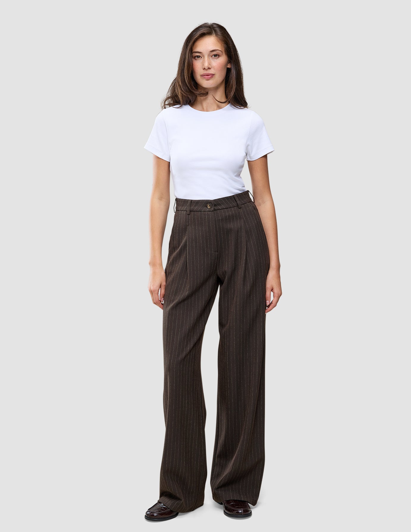 En person iført en hvid T-shirt og et par Serene Pants Wide Walnut Herringbone, som er mørkebrune bukser med brede ben og herringbone-mønster.