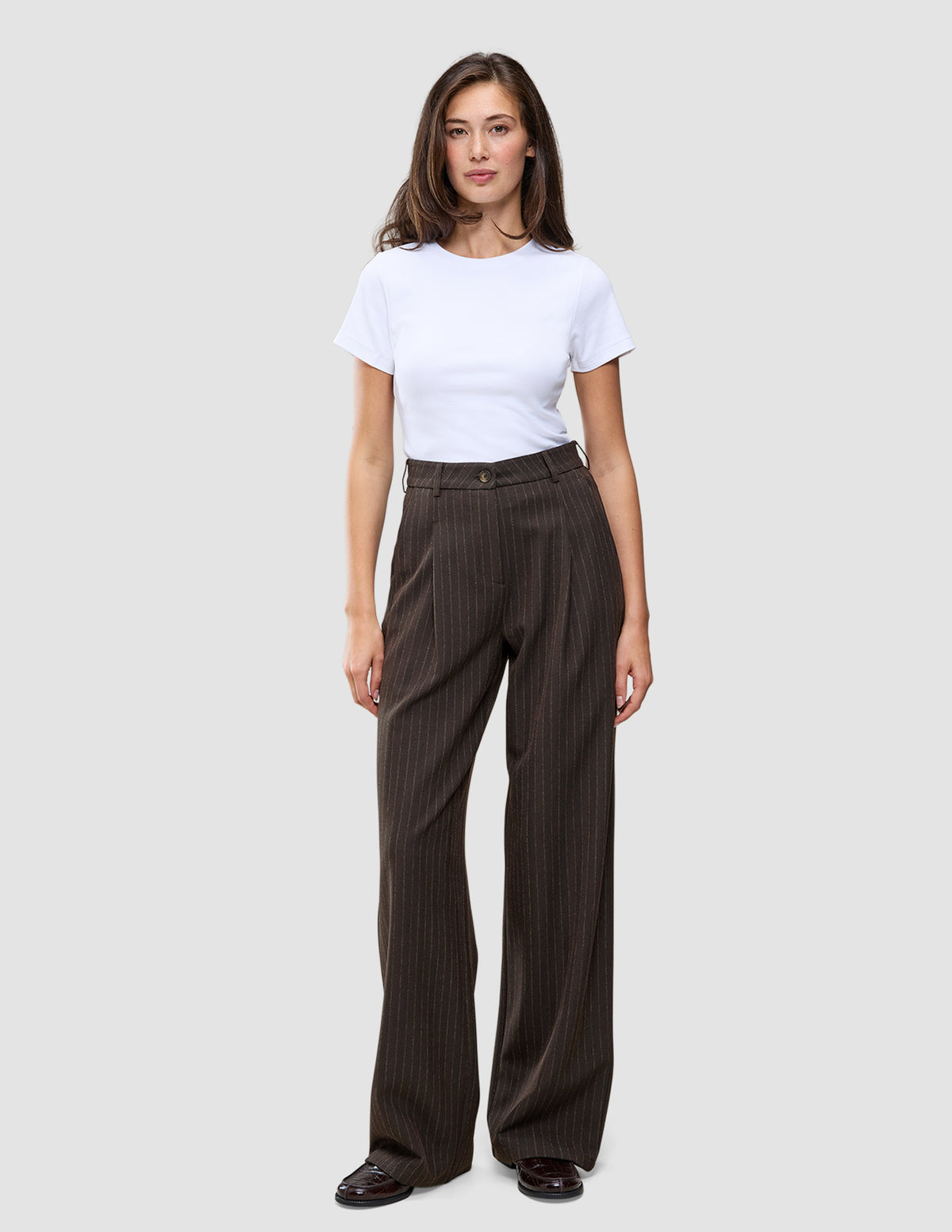 En person iført en hvid T-shirt og et par Serene Pants Wide Walnut Herringbone, som er mørkebrune bukser med brede ben og herringbone-mønster.