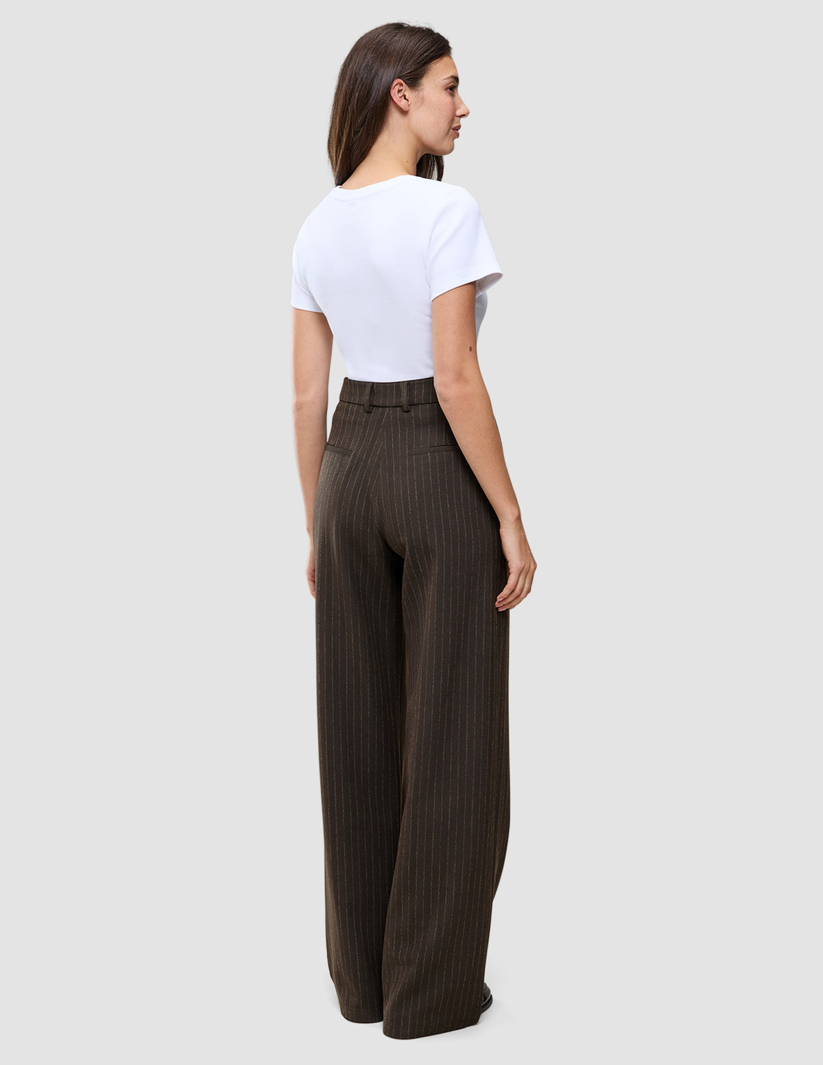 En person set bagfra, iført en hvid T-shirt og mørkebrune Serene Pants Wide Walnut Herringbone-bukser med høj talje og brede ben.