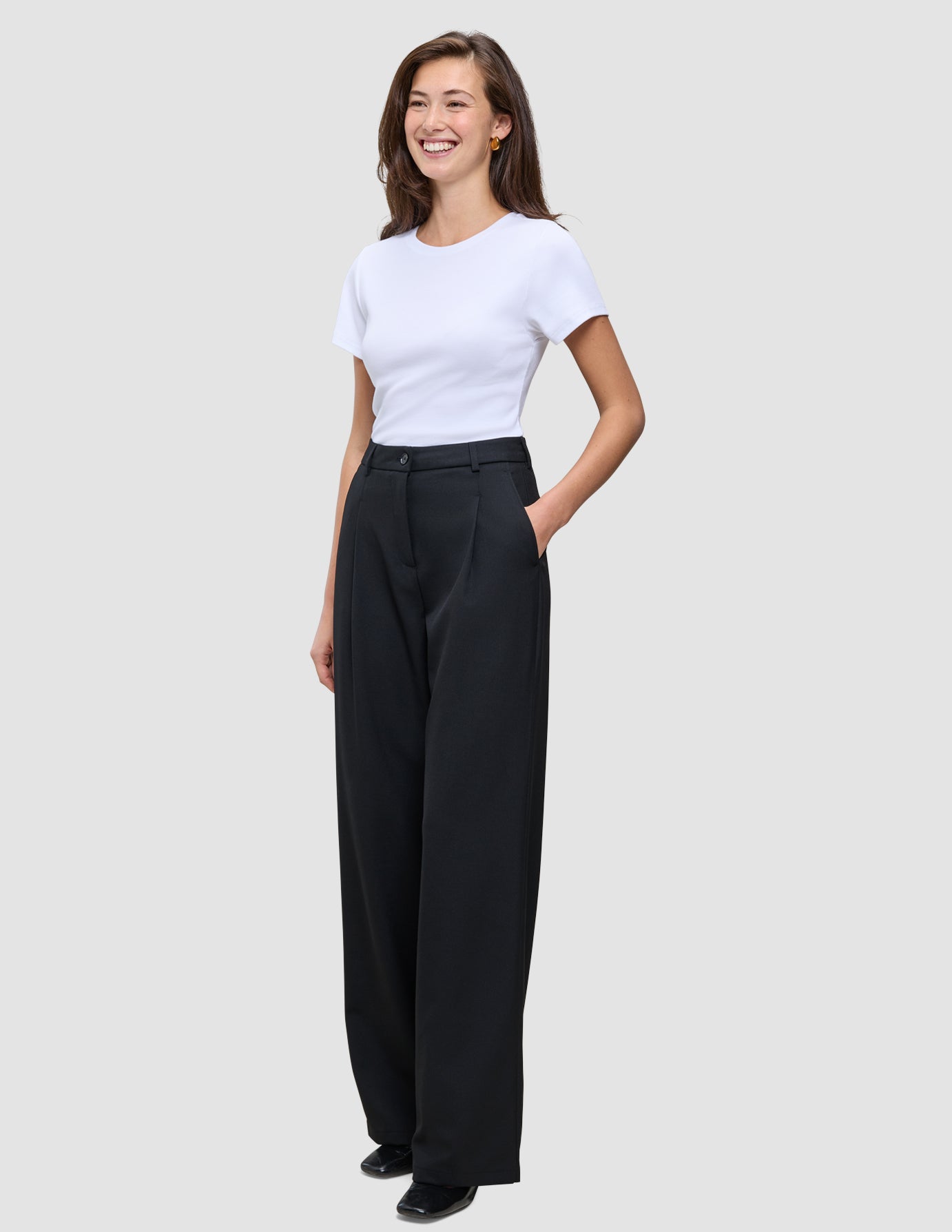 En smilende kvinde har en hvid T-shirt på og et par Serene Pants Wide Black med brede ben, og hun har en hånd i lommen.