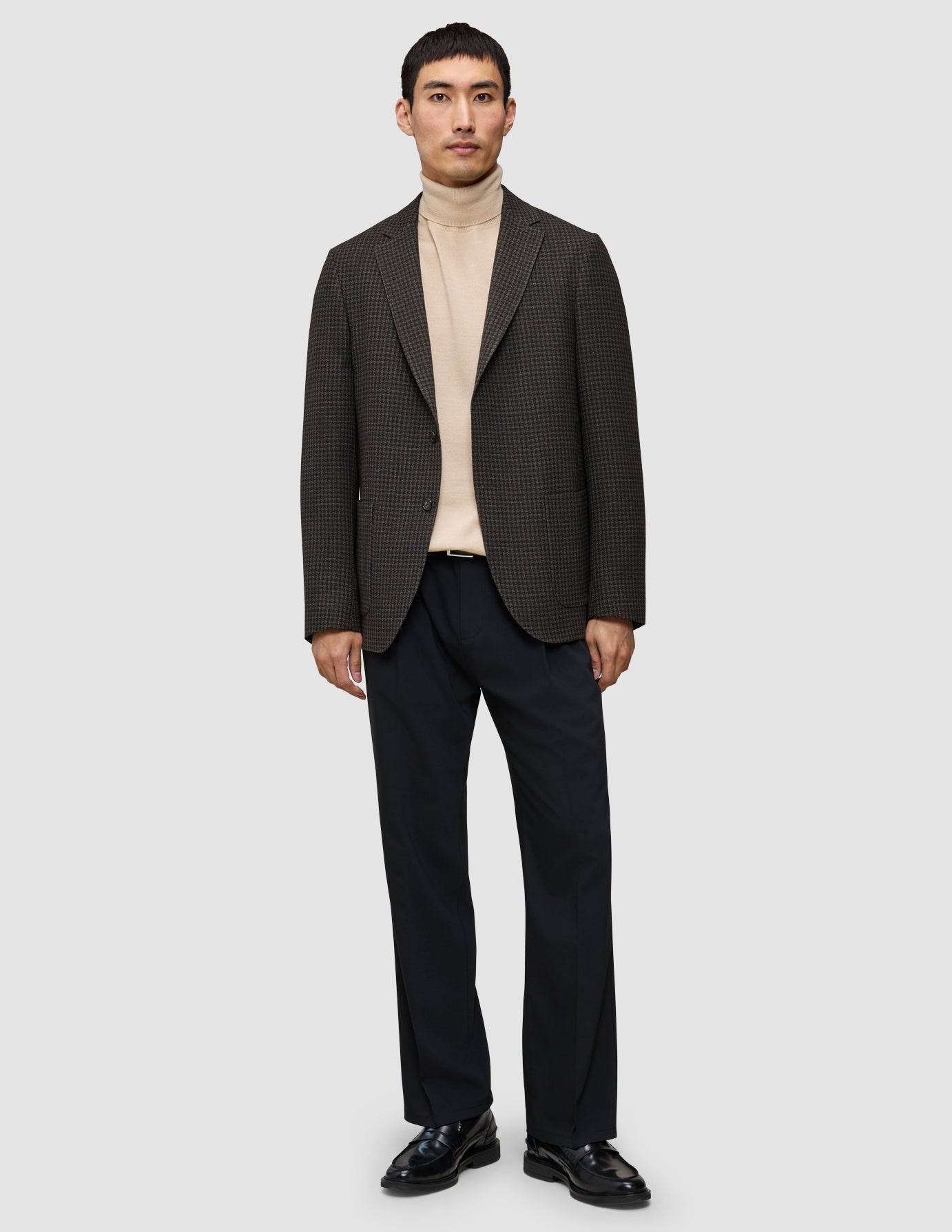 En mand i et look med strik og blazer bestående af en merino-trøje med rund hals, en mørk uldblazer med mønster, sorte bukser og sorte loafers.