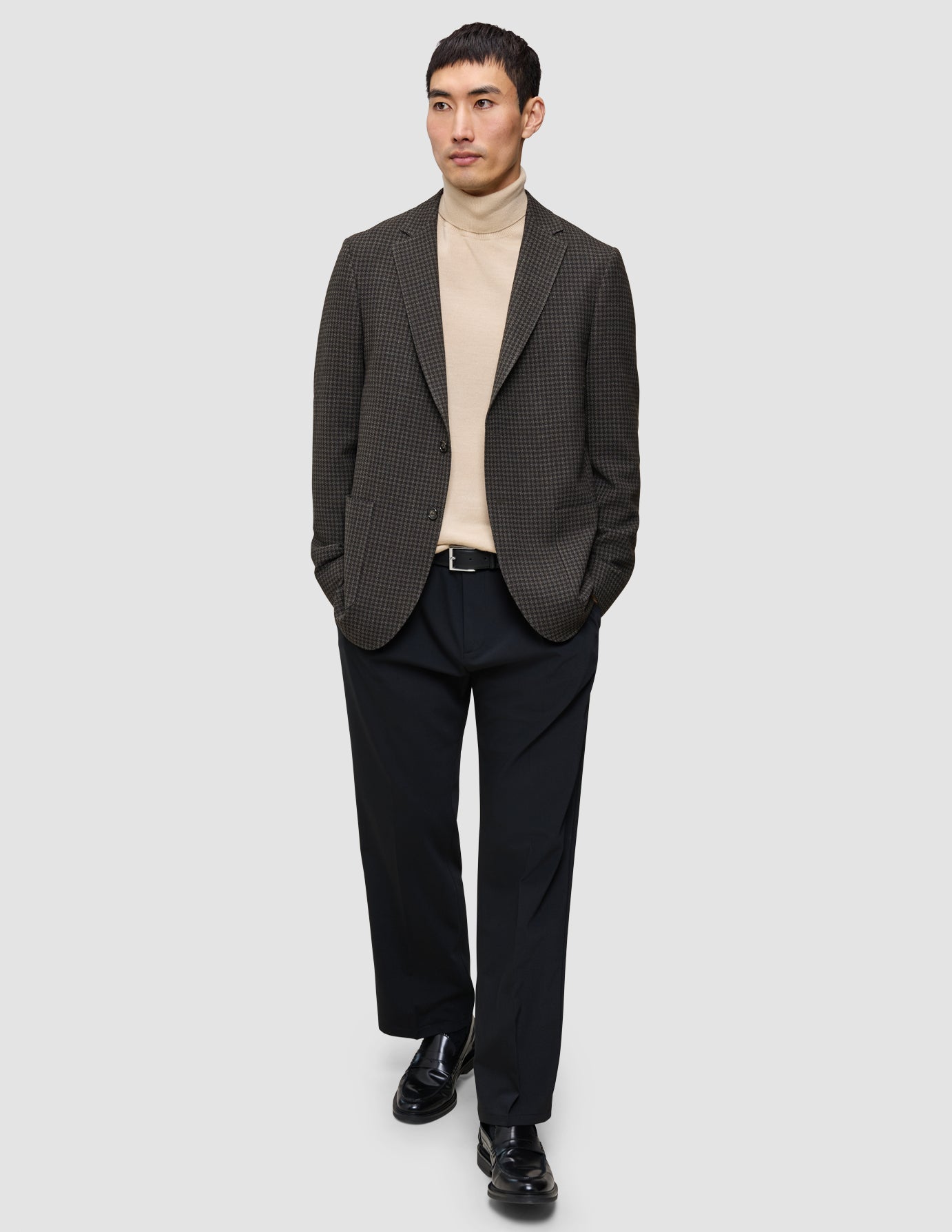 En person i et look med strik og blazer, iført en beige Premium Merino-trøje med rund hals, en mørk mønstret blazer og sorte loafers.